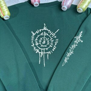 K&ouml;nnte beinhalten: Ein dunkelgr&uuml;nes Sweatshirt mit wei&szlig;en Stickereien. Die Vorderseite hat ein kreisf&ouml;rmiges Design mit Text, und der &Auml;rmel hat ein Schwert- und Blumendesign. Das Sweatshirt ist ein Rundhals-Stil.