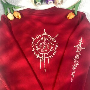 K&ouml;nnte beinhalten: Ein rotes Sweatshirt mit wei&szlig;en Stickereien. Auf der Vorderseite befindet sich ein kreisf&ouml;rmiges Emblem mit komplizierten Details, w&auml;hrend der &Auml;rmel ein Schwertdesign zeigt. Das Sweatshirt ist ein Rundhals-Stil und scheint aus einem weichen Material zu sein.