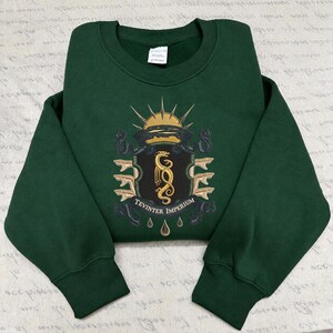 Könnte beinhalten: Ein dunkelwaldgrüner Rundhals-Pullover mit einem zentralen gestickten Wappen. Das Design zeigt einen goldenen Drachen, eine aufgehende Sonne und den Schriftzug Tevinter Imperium auf einem dunklen Banner, flankiert von stilisierten Schlangenköpfen.