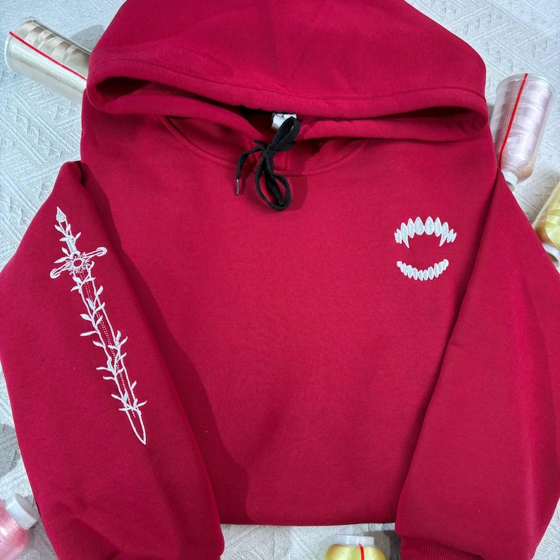 K&ouml;nnte beinhalten: Ein weinroter Kapuzenpullover mit Kordelzug und wei&szlig;en Stickereien. Der linke &Auml;rmel zeigt ein Schwertdesign, w&auml;hrend die Brust ein zahniges Grinsen zeigt. Der Hoodie ist aus weichem Material.