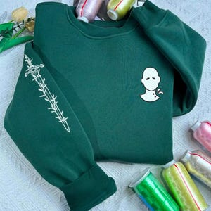 Può includere: Una felpa verde scuro con una grafica bianca di un volto sul petto e un disegno di spada bianca sulla manica sinistra. La felpa è in stile casual ed è realizzata in un materiale morbido.