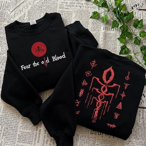 May include: Two black crewneck sweatshirts featuring red embroidery. One sweatshirt displays a red circle with a spider-like design and the text "Fear the old Blood". The other sweatshirt features a large, intricate red symbol surrounded by smaller red icons.