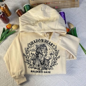 May include: Cream-colored hooded sweatshirt featuring a black graphic of a woman's portrait with the text "SHADOWHEART" and "BALDUR'S GATE" in a circular design. The sweatshirt has a drawstring hood and ribbed cuffs.