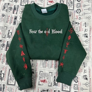 May include: Dark green sweatshirt with the text "Fear the old Blood" in white and red embroidered symbols on the sleeves. The sweatshirt has a crew neck and ribbed cuffs.