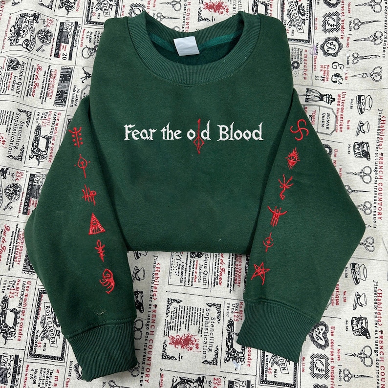 May include: Dark green sweatshirt with the text "Fear the old Blood" in white and red embroidered symbols on the sleeves. The sweatshirt has a crew neck and ribbed cuffs.