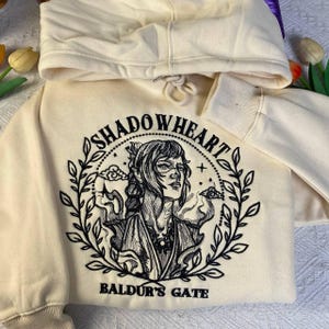 May include: Cream-colored hoodie with a black embroidered design featuring the text "SHADOWHEART" and "BALDUR'S GATE" around a portrait of a woman. The design is framed by a wreath of leaves.