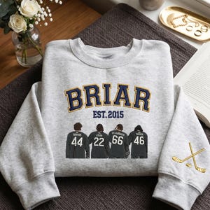 Op de afbeelding: Een lichtgrijs sweatshirt met het woord "BRIAR" in marineblauw en goud, met "EST. 2015" eronder. Het sweatshirt heeft vier figuren met rugnummers en namen, en gekruiste hockeysticks op de mouw.