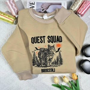 Könnte beinhalten: Ein beiger Pullover mit einem schwarz-weißen Motiv einer Katze mit Rucksack und dem Text "QUEST SQUAD BROCCOLI".