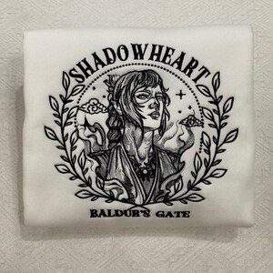 May include: White sweatshirt featuring a black illustration of a woman surrounded by a wreath. The text "SHADOWHEART" arches above the illustration, with "BALDUR'S GATE" below. The design is centered on the folded sweatshirt.
