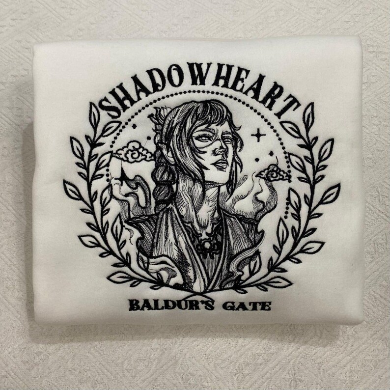 May include: White sweatshirt featuring a black illustration of a woman surrounded by a wreath. The text "SHADOWHEART" arches above the illustration, with "BALDUR'S GATE" below. The design is centered on the folded sweatshirt.