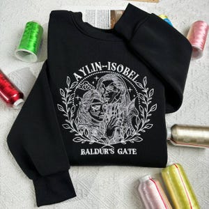 Puede incluir: Sudadera negra con bordado blanco de dos figuras abrazándose, rodeadas de una corona y el texto "Aelyn-Isobel Baldur's Gate".