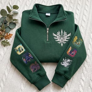 Puede incluir: Una sudadera verde oscuro con cremallera de un cuarto, un emblema blanco bordado en el pecho e ilustraciones coloridas de criaturas impresas en ambas mangas. La prenda tiene cuello alto, una cremallera metálica plateada y puños acanalados en las muñecas.
