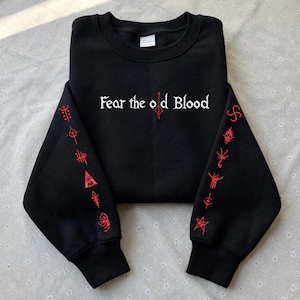 以下が含まれることがあります： 「Fear the old Blood」の白文字がプリントされた黒いスウェットシャツ。袖には赤い刺繍のシンボルが施されています。デザインを際立たせるように、衣服はきれいに折りたたまれています。
