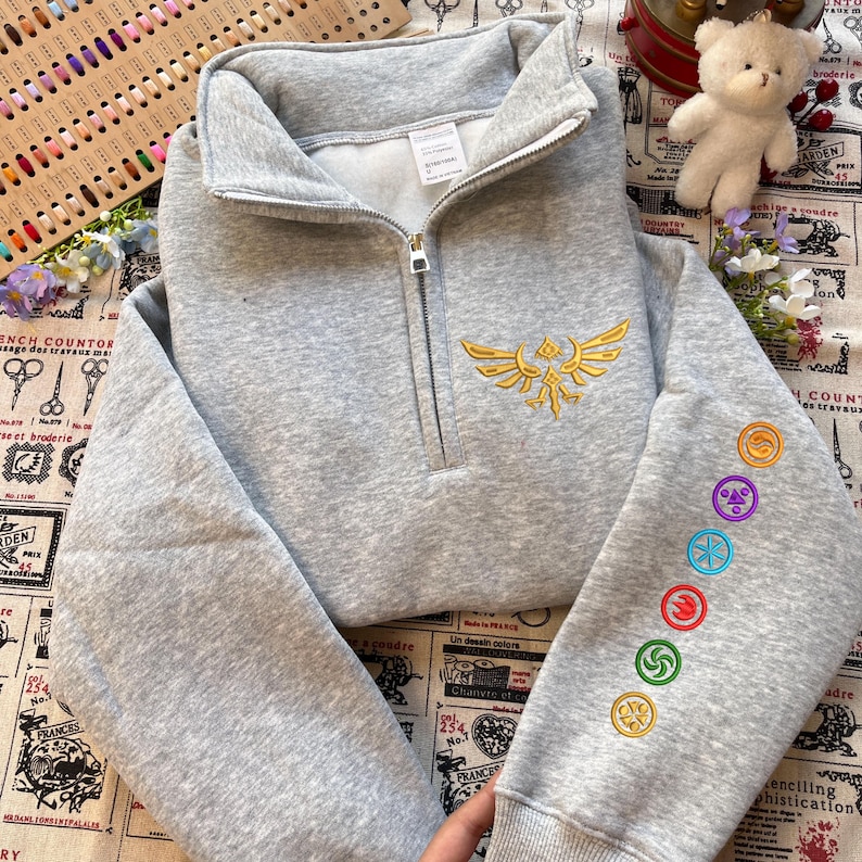 May include: A gray quarter-zip sweatshirt with a gold embroidered emblem on the chest and colorful circular designs on the sleeve. The sweatshirt has a high collar and a zipper closure.