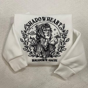 May include: White sweatshirt with the text "SHADOWHEART" above an illustration of a woman's face, surrounded by a laurel wreath. The text "BALDUR'S GATE" is below the illustration. The sleeves are cuffed.