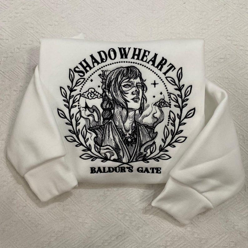 May include: White sweatshirt with the text "SHADOWHEART" above an illustration of a woman's face, surrounded by a laurel wreath. The text "BALDUR'S GATE" is below the illustration. The sleeves are cuffed.