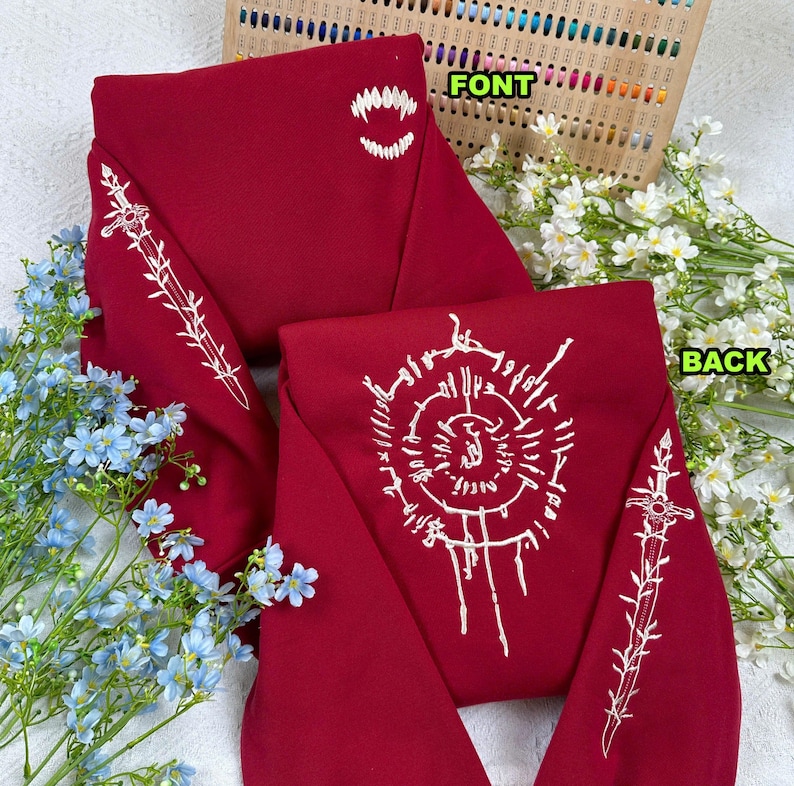 K&ouml;nnte beinhalten: Ein tiefrotes Sweatshirt mit wei&szlig;en Stickereien. Die Vorderseite zeigt ein Gebiss, w&auml;hrend die R&uuml;ckseite ein kompliziertes kreisf&ouml;rmiges Muster zeigt. Die &Auml;rmel haben ein Schwertdesign. Das Sweatshirt ist von blauen und wei&szlig;en Blumen umgeben.