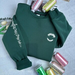 K&ouml;nnte beinhalten: Ein dunkelgr&uuml;nes Sweatshirt mit wei&szlig;en Stickereien. Der linke &Auml;rmel zeigt ein Schwertdesign, w&auml;hrend die Vorderseite ein stilisiertes Fangzahn-Design aufweist. Das Sweatshirt ist aus weichem Material und hat einen Rundhalsausschnitt.