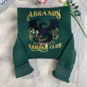 以下が含まれることがあります： ゴールドの文字で「ABRAXOS GARDEN CLUB」と書かれた濃い緑色のスウェットシャツ。中央には黒いドラゴンが刺繍され、花のディテールで囲まれています。スウェットシャツはリブ編みの袖口とクルーネックです。