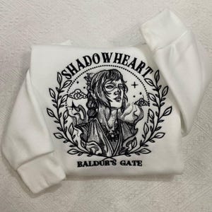 May include: White sweatshirt featuring a black and white illustration of a character with the text "SHADOWHEART" above the image and "BALDUR'S GATE" below. The design is surrounded by a wreath of leaves.