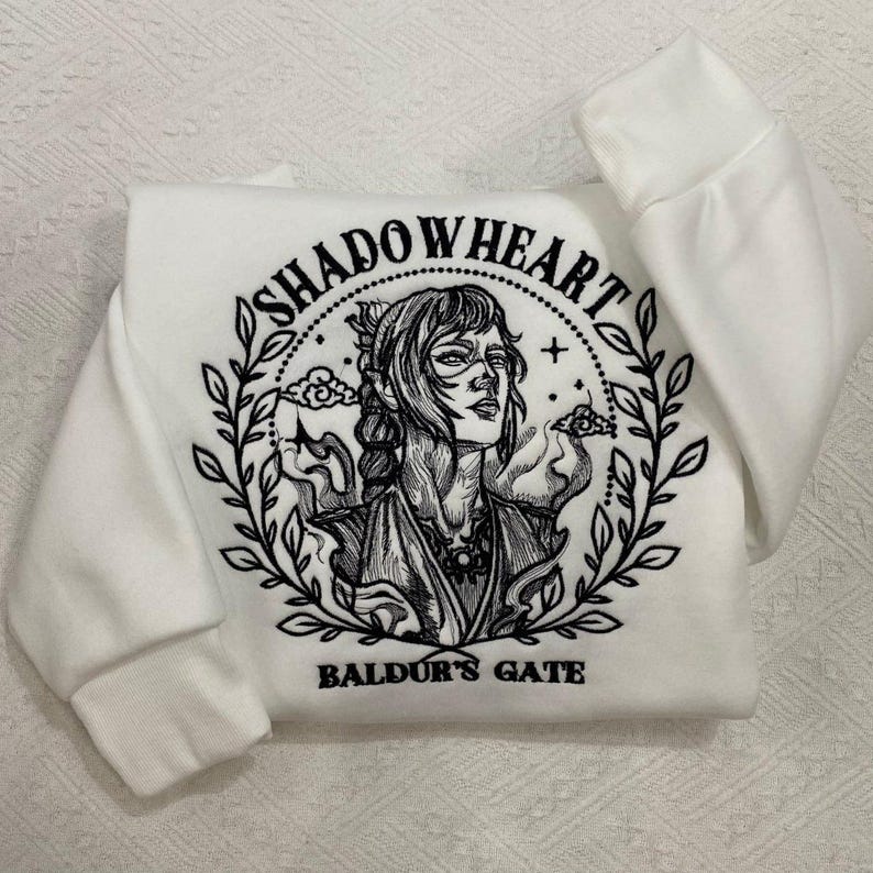 May include: White sweatshirt featuring a black and white illustration of a character with the text "SHADOWHEART" above the image and "BALDUR'S GATE" below. The design is surrounded by a wreath of leaves.