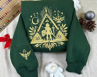 May include: A dark green sweatshirt with a gold embroidered design featuring a triangle, a figure with a sword and shield, and other decorative elements. The sleeve has a gold emblem. The sweatshirt is a cozy, casual garment.
