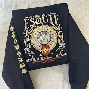 May include: Black sweatshirt with gold embroidery. The design includes the word "ESQUIF", a sun-like emblem, and the text "MASTER OF SKY, SEA... AND NAPS." The left sleeve has additional gold text. The sweatshirt is folded.