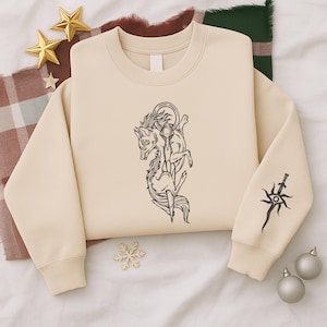 May include: Cream-colored sweatshirt with a black graphic of a wolf-like creature and a sword design on the sleeve. The sweatshirt has a crew neck and long sleeves. The design is centered on the front.