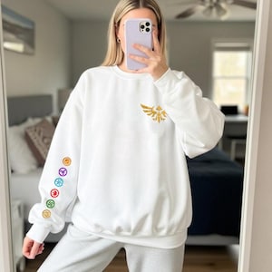 May include: White sweatshirt with a gold emblem on the chest and colorful circular designs on the left sleeve. The sweatshirt is paired with gray sweatpants. The image is taken in front of a mirror.