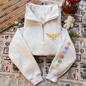 May include: A light gray cropped sweatshirt with a quarter-zip closure and a stand-up collar. The front features a gold embroidered emblem, and the left sleeve displays colorful embroidered symbols. The sweatshirt is made of a soft, comfortable material.