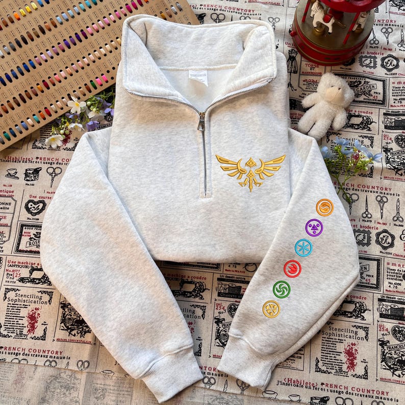 May include: A light gray cropped sweatshirt with a quarter-zip closure and a stand-up collar. The front features a gold embroidered emblem, and the left sleeve displays colorful embroidered symbols. The sweatshirt is made of a soft, comfortable material.