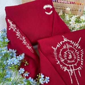 K&ouml;nnte beinhalten: Gefalteter roter Stoff mit wei&szlig;en Stickereien. Ein Abschnitt zeigt ein Schwertdesign, ein anderer Z&auml;hne und ein dritter ein kreisf&ouml;rmiges Muster mit Text. Blaue und wei&szlig;e Blumen umgeben den Stoff.