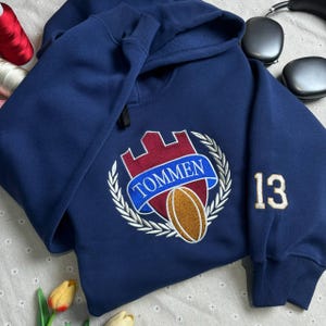 Puede incluir: Una sudadera con capucha azul marino con un escudo bordado en blanco y plateado que presenta un castillo, una pelota de rugby y el texto "TOMMEN". El número "13" está bordado en la manga.
