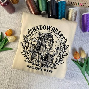 May include: Cream-colored sweatshirt featuring a detailed black and white embroidered design of a character from Baldur's Gate. The design includes the text "SHADOWHEART" and "BALDUR'S GATE" in a circular arrangement around the character's portrait. The sweatshirt is surrounded by spools of thread.