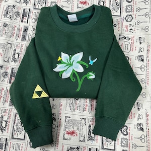 May include: A dark green sweatshirt with a white and light blue floral embroidery design on the front. A small yellow triangle is on the left sleeve. A small blue bird is embroidered near the flower.