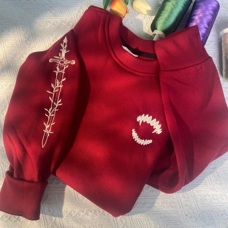 K&ouml;nnte beinhalten: Ein weinrotes Sweatshirt mit wei&szlig;en Stickereien. Eine Seite zeigt ein Schwertdesign, die andere ein l&auml;chelndes Munddesign. Das Sweatshirt hat einen Rundhalsausschnitt und lange &Auml;rmel mit gerippten B&uuml;ndchen.