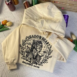 May include: Cream-colored hoodie featuring a black and white illustration of a woman with the text "SHADOWHEART" and "BALDUR'S GATE". The hoodie has a drawstring hood and ribbed cuffs.