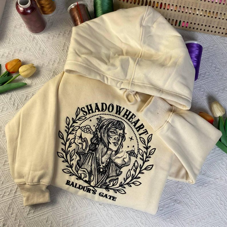 May include: Cream-colored hoodie featuring a black and white illustration of a woman with the text "SHADOWHEART" and "BALDUR'S GATE". The hoodie has a drawstring hood and ribbed cuffs.
