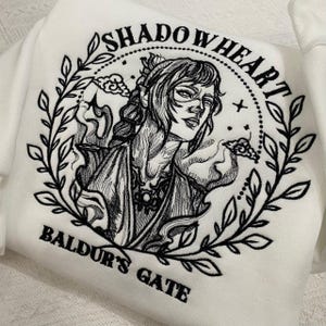 May include: White sweatshirt with a black embroidered design featuring a woman's portrait, surrounded by leaves and the text "SHADOWHEART" and "BALDUR'S GATE". The design is detailed and artistic.
