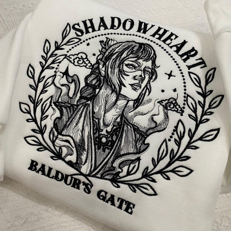 May include: White sweatshirt with a black embroidered design featuring a woman's portrait, surrounded by leaves and the text "SHADOWHEART" and "BALDUR'S GATE". The design is detailed and artistic.