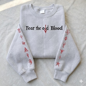 May include: A light gray sweatshirt with the text "Fear the old Blood" in black and red. The sleeves feature red symbols. The sweatshirt has a crew neck and long sleeves.