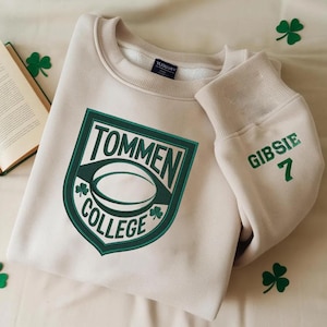 May include: A beige sweatshirt with a green embroidered patch that says "Tommen College" and a rugby ball. The sleeve has the name "Gibsie 7" embroidered in green.