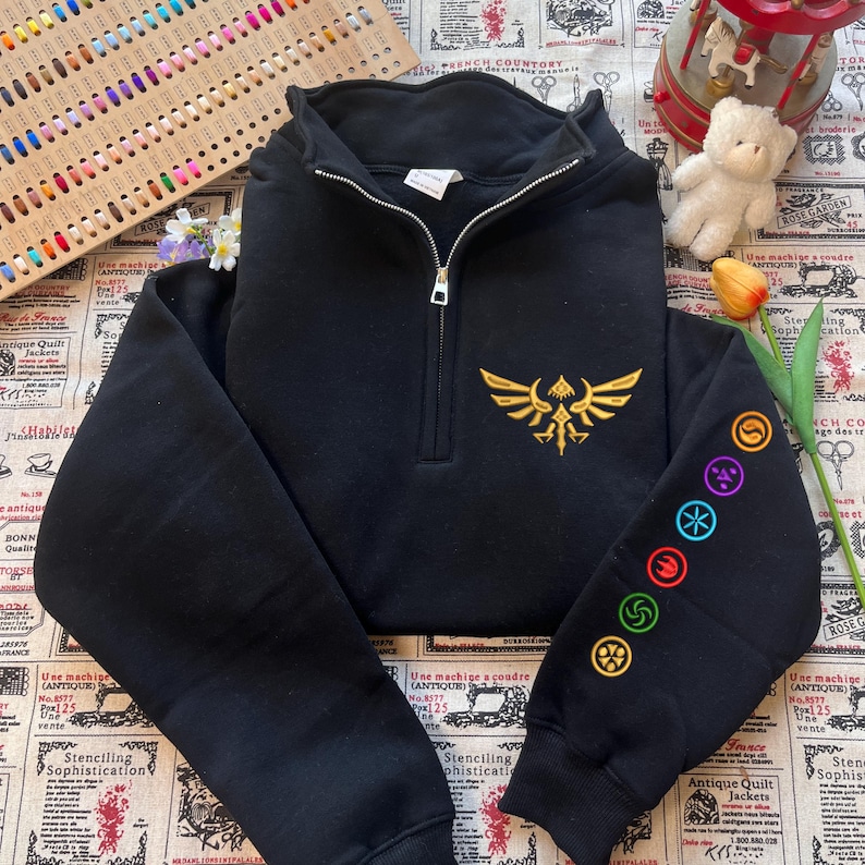 May include: Black quarter-zip sweatshirt with a gold embroidered emblem on the chest and colorful symbols on the left sleeve. The sweatshirt has a zipper closure and long sleeves. The design is inspired by a popular video game.