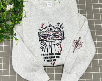 May include: A light gray heathered crewneck sweatshirt featuring a black and red embroidered graphic of eyes within a rectangular frame, surrounded by abstract lines and droplets. The text reads, I'M SO MUCH MORE THAN WHAT YOU MADE ME.