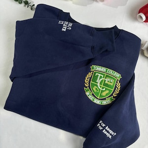 Puede incluir: Una sudadera azul marino con un escudo bordado en verde y dorado con el texto "Tommen College" y "In Hoc Signo Vinces". La sudadera también tiene un texto bordado en blanco en la manga que dice "For keeps? For keeps."