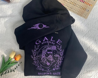 May include: Black hooded sweatshirt with purple graphics. The front features a detailed illustration of a man with the text "GALE APPROVES" and "BALDUR'S GATE". The left sleeve has a purple design. The sweatshirt is folded on a white surface.