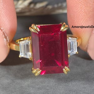 May include: A gold ring featuring a large, rectangular, deep red gemstone, flanked by two smaller, rectangular, clear gemstones. The ring is held up against a gray background. The text "AmarajewelsDesign" is visible.
