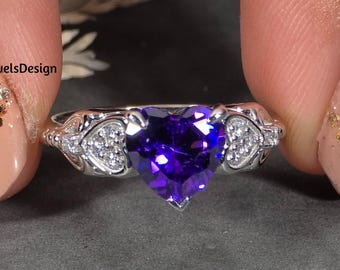 May include: A silver ring featuring a heart-shaped, deep purple gemstone. The ring has a delicate design with small, clear accent stones on either side of the heart. The ring is held between two fingers.