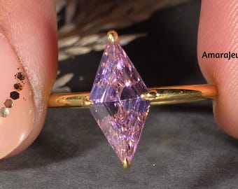 May include: A gold ring featuring a kite-shaped, light purple gemstone. The ring is held between two fingers, showcasing the intricate facets of the gemstone. The ring's band is thin and gold, and the gemstone is set with gold prongs. The text "AmarajewelsDesign" is visible.