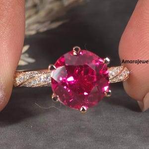 以下が含まれることがあります： 大きな丸い鮮やかなピンク色の宝石が付いたローズゴールドの指輪。バンドは、小さく透明なアクセントストーンを備えた繊細なツイストデザインが特徴です。指輪は、暗い背景に対して2本の指の間に保持されています。